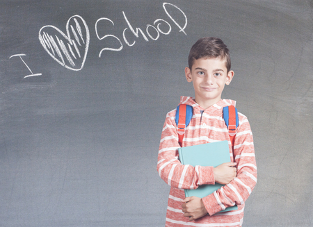 Smiling school boy with backpack holding a book in front of a blackboard