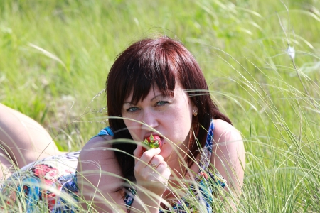 Beautiful brunette is eating a strawberry while lying in the grassの写真素材