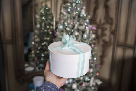 Against the background of the New Year's decorated fir-tree the female hand holds a white gift with...