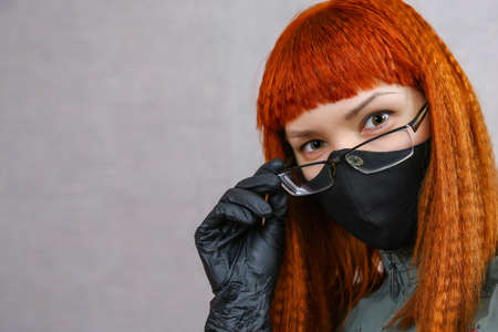 a red-haired girl in a black cloth mask, black medical gloves and glasses looks into the frame. a young beautiful woman holds her glasses in her hand. covid-19.の写真素材