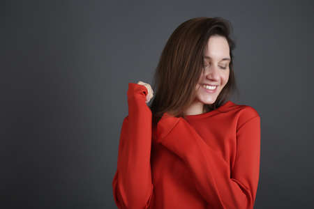 smiling young woman with closed eyes in a red sweater smiles on a gray background.