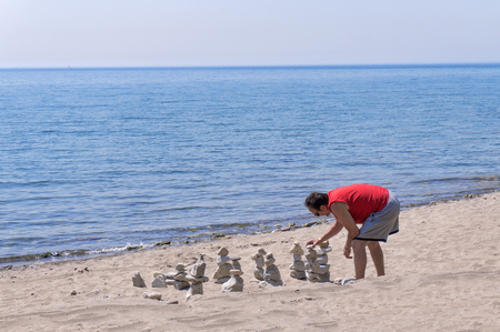 A man in red t-shirt does zen practice by balancing and stacking stones at beach in early summer.のeditorial素材