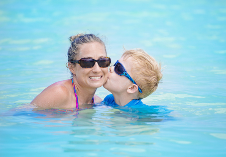 Mother and son playing together in an outdoor swimming pool. Boy kissing his smiling mom in the water. Fun time togetherの写真素材