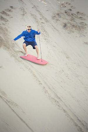 Thrill-seeking mid adult male riding a board down a sand dune hill having fun playing outdoors while on vacation.の写真素材