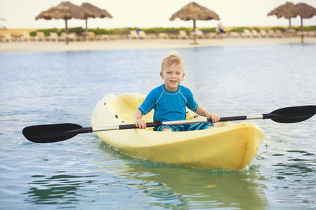 Young active Boy paddling an ocean kayak at the beach while on a tropical vacation. Child having fun a scenic beach resort with lounge chairs and cabanas in the backgroundの写真素材