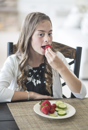 Cute girl sitting at the dinner table eating a healthy plate of fruits and vegetables. She is developing good eating habits eating strawberries and cucumberの写真素材