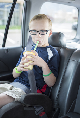 Cute boy sitting in a booster seat on a long car ride drinking a smoothie while safely strapped in a car seat. He is wearing his seatbelt and enjoying a drink.の写真素材