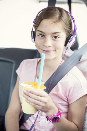 Cute smiling girl on a long car ride listening to music and drinking while safely strapped in a car seatの写真素材
