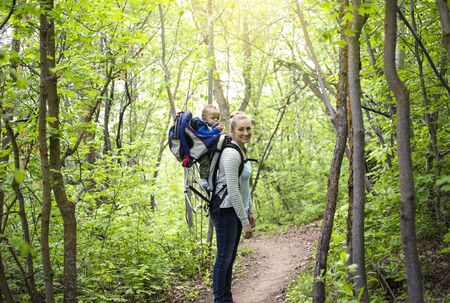 Young mother and her son take a hike together in the mountains on a beautiful summer evening. Cute young baby son riding on moms shoulders in a backpackの写真素材