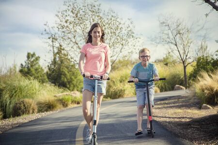 Two smiling kids having fun on a scooter ride outdoors on a paved pathway on a sunny day. Active children playing together outdoorsの写真素材