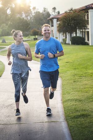 Beautiful woman and mature man jogging together outdoors along a pathway at a city park. Happy and smiling as they run along the path during sunset on a warm summer dayの写真素材