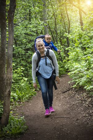 A young mother and her son take a hike together in the mountains on a beautiful summer evening. Cute young baby son riding on moms shoulders in a backpackの写真素材