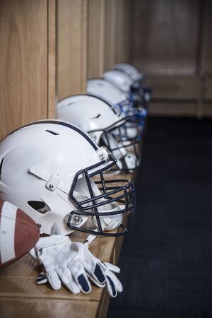 Close up view of a row of American football helmets sitting in a locker room before an football game. Gloves and a football also sitting on the bench in the locker roomの写真素材