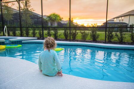Rear view of a child sitting on the edge of a backyard swimming pool. Concept photo of swimming pool danger and child water safety.の写真素材
