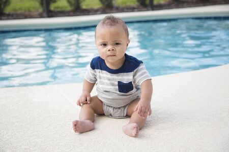 Cute baby boy sitting dangerously on the edge of a swimming. Concept photo of swimming pool danger and water child safety. An unattended young baby in danger of falling in and drowningの写真素材