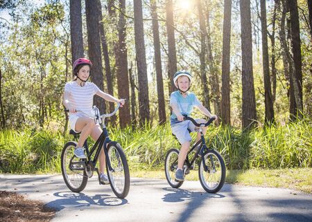 Two kids enjoying a fun bicycle ride together on a bike path in a green forest outdoors. Smiling and enjoying nature on a sunny warm day. Exercising and being active during the summerの写真素材