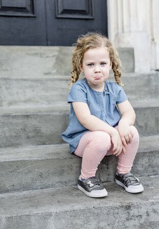 Emotional portrait of a sad, unhappy little girl with blond curly hair sitting on the front steps of her home in an urban setting. Cute expression and adorable face. child behavior concept photoの写真素材