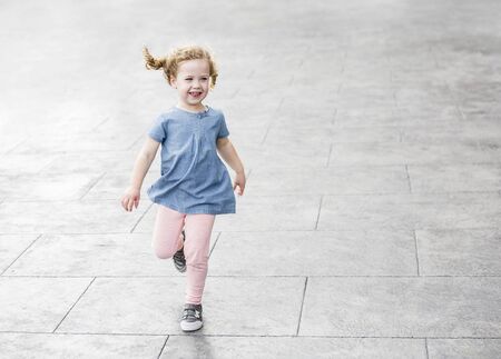 portrait of a happy energetic little girl with blond curly hair laughing as she runs around outdoors on a paved walk way. Carefree, Joy, fun and Happiness concept photoの写真素材