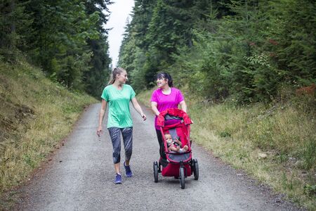 Two women in their 30s walking and talking together on a trail with pushing a baby strollerの写真素材