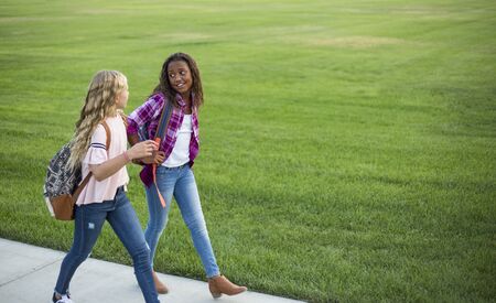 Two diverse school kids walking and talking together on the way to school. Back to school photo of diverse girls wearing backpacks in the school yardの写真素材