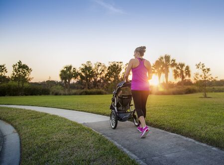 Beautiful, fit women walking and jogging outdoors along a paved sidewalk in a park pushing a stroller at sunsetの写真素材