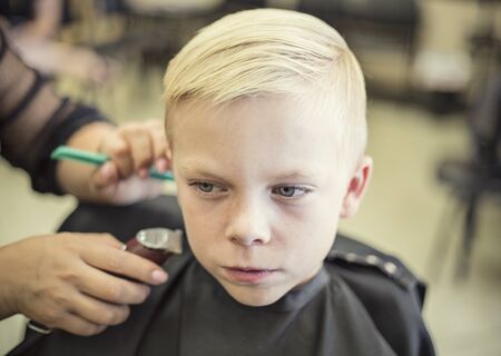 A cute young blonde boy getting his hair cut at a beauty salon or barbershop by a female hair stylistの写真素材
