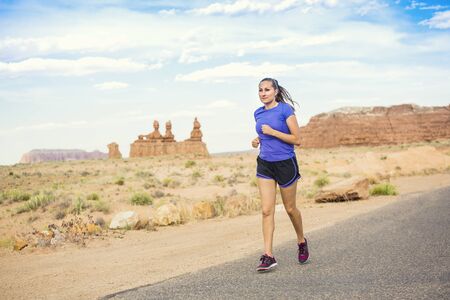 Beautiful fit woman in her 30s running on a paved trail in a scenic desert park. The healthy woman stays fit even on vacation with rocky sandstone cliffs in the backgroundの写真素材