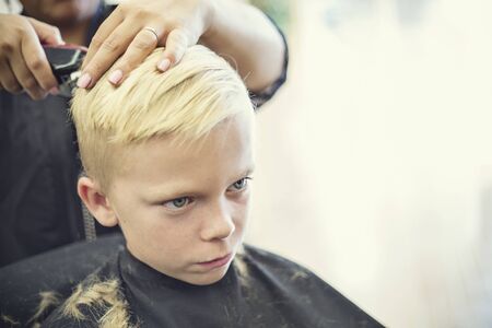 A cute young blonde boy getting his hair cut at a beauty salon or barbershop by a female hair stylistの写真素材