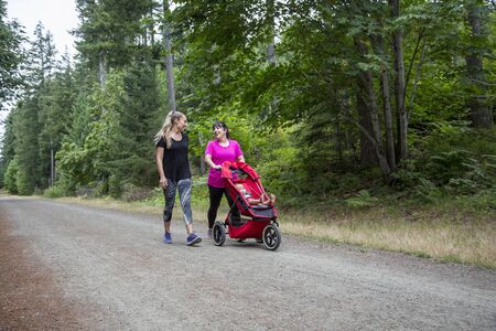 Two women in their 30s walking and talking together on a trail with pushing a baby stroller. Walking along a beautiful scenic nature trail.の写真素材