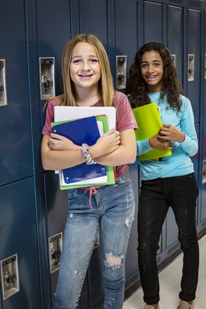 Portrait of two diverse female Junior High school Student standing in a school hallway. Smiling school girls looking and holding their books during breaks in the dayの写真素材