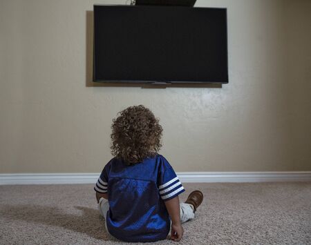 Rear view of a young child watching Television while sitting on the floor of his home. Selective focus on the back of the curly-haired diverse little boy watching sports on TVの写真素材