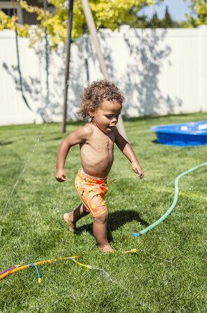 Smiling Cute little boy splashing through the sprinklers in the backyard. African American boy...