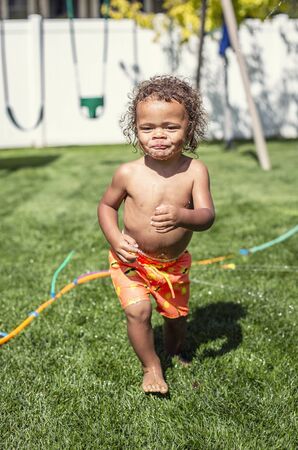Laughing Cute little boy splashing through the sprinklers in the backyard. African American boy playing in the outdoors on a warm summer dayの写真素材