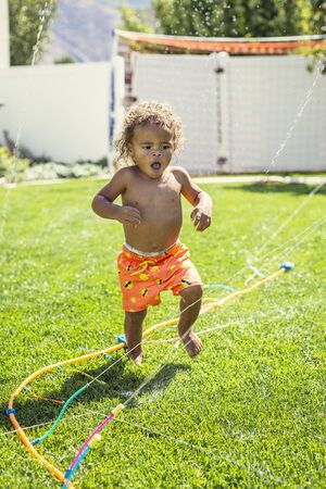 Smiling Cute little boy splashing through the sprinklers in the backyard. African American boy playing in the outdoors on a warm summer dayの写真素材