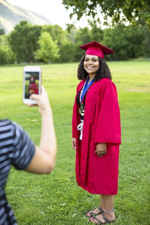Taking a smartphone photo of a recent high school graduate in her cap and gown. Full length candid photo of a cute smiling diverse girl of African descent.の写真素材