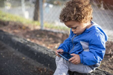 Young diverse boy using his parent`s smartphone at a park outdoors. Sitting and looking into a...