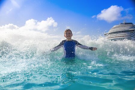 Happy Boy playing in the ocean waves while on a Cruise vacation. Aqua blue Caribbean sea water splashing the boy from behind on a warm day in the islands.の写真素材