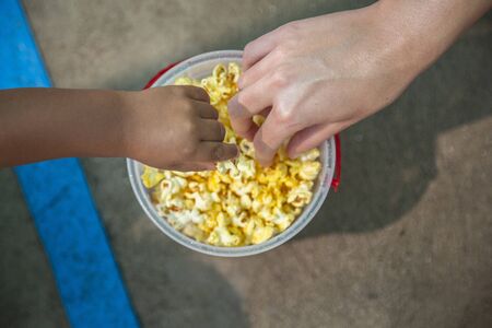 Close up photo of two hands reaching for buttered popcorn in a bucket at an amusement parkの写真素材