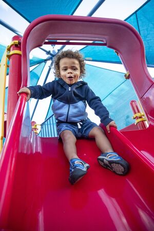 Happy cute young diverse boy with enjoying a ride down playground slide at an outdoor park. Excited and fun expressionの写真素材