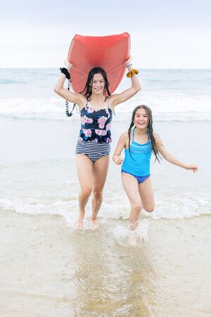 Full length photo of two cute teen girls running on a beautiful beach carrying a bodyboard. Having fun in the sun while on a family beach vacation.の写真素材