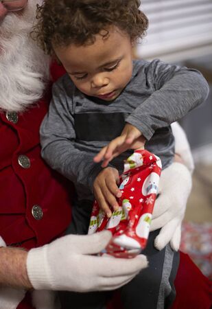 Close up view of a Young bi-racial child opening a Christmas present on Santa`s lap. Santa Clause giving a cute little boy a present that he is unwrapping with excitementの写真素材