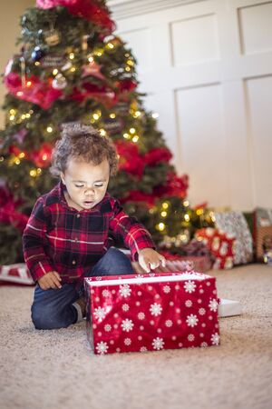 Cute little diverse boy opening a magical Christmas present in front of a Christmas tree. Excited expression on the child's face as he looks inside the boxの写真素材