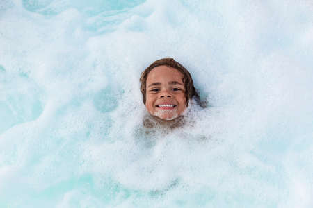 Cute smiling boy playing in the bubbles of a hot tub spa or bath tub. Fun, happy expression of a cute African American boy playing in the foamy waterの写真素材