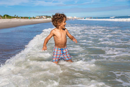 Cute young mixed race boy running and playing on the beach while on a family vacation. Playing in the ocean waves having funの写真素材
