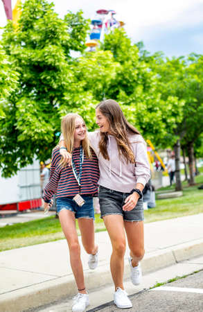 Two smiling teenage girls walking together at an outdoor carnival. Best friends having fun during summer vacation. Friendship and happiness.の写真素材