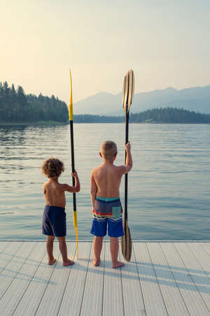 Two boys on the edge of the dock of a beautiful scenic lake during their summer vacation. Ready to paddle in the waterの写真素材