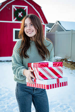Beautiful young woman walking and delivering gifts during the Christmas season. Walking by a red barn with a Christmas wreath and looking forward to giving during the holiday seasonの写真素材