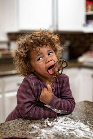 Cute mixed-race boy licking the blender beater from a bowl of brownie cake batter as he makes a mess in the kitchen. Child having fun at home baking and cookingの写真素材