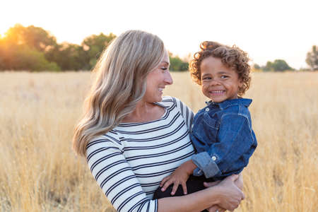 Beautiful happy Mother holding her adorable diverse son in the outdoor sunlight. Sun rays shining through as the mom shows love and affection to her sonの写真素材