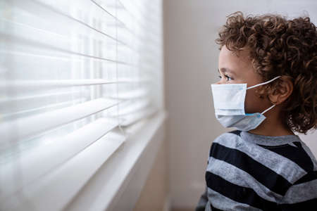 A young boy looking out the window wearing a protective facemark while seeking protectionの写真素材